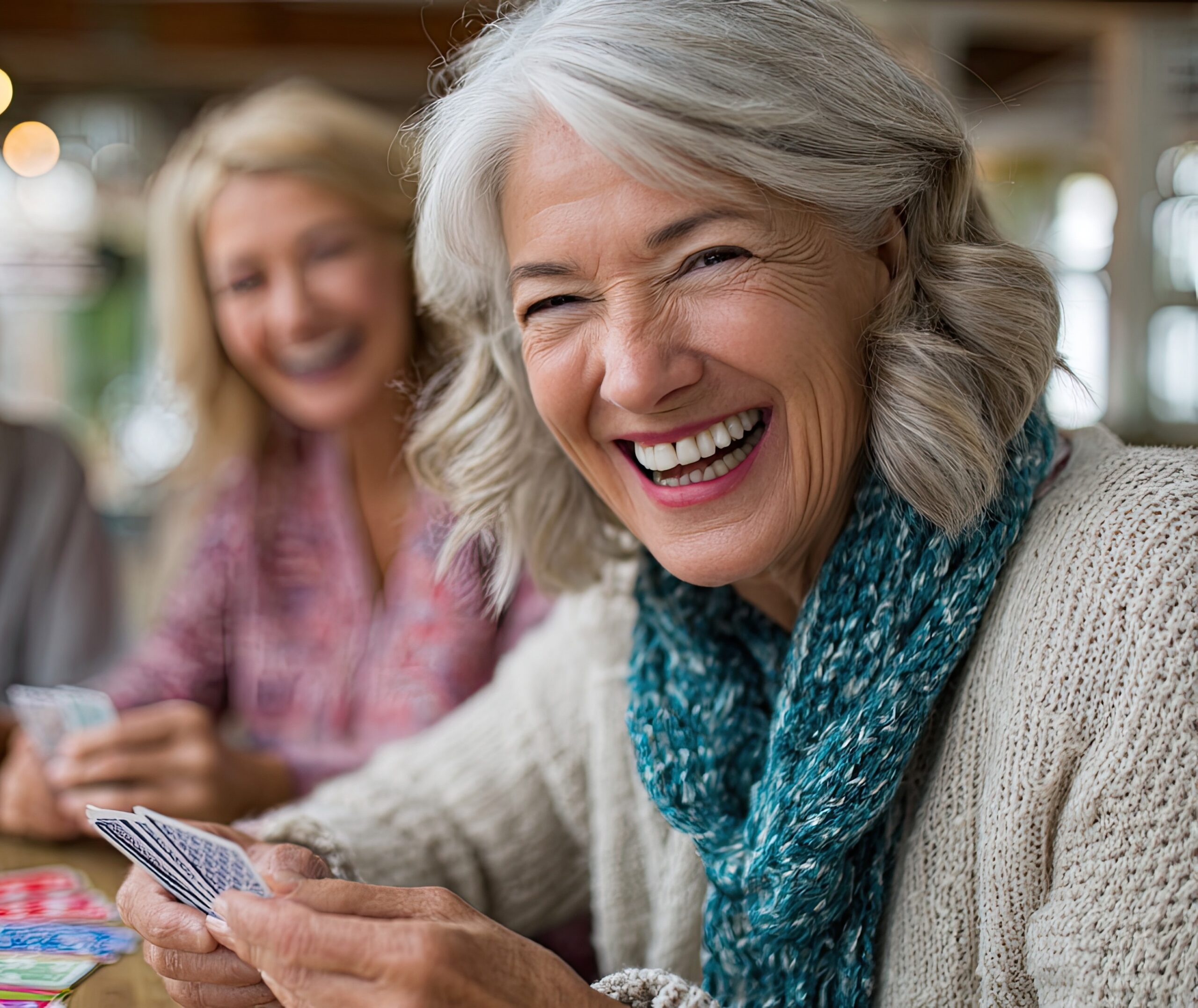 An older woman with gray hair and a teal scarf smiles and laughs while playing cards with a friend in a cozy, well-lit senior living community in Hilliard, Ohio. Another woman in the background also holds cards and is smiling.