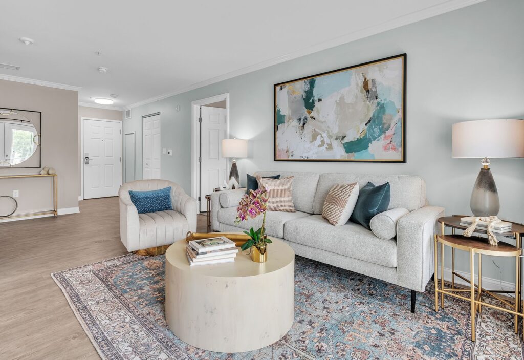 Modern living room with a light gray sofa, armchair, abstract wall art, and a patterned rug exudes luxury. A round coffee table with books and a plant sits centerstage. A desk with mirror is near the entry door in this inviting Hilliard, Ohio space.