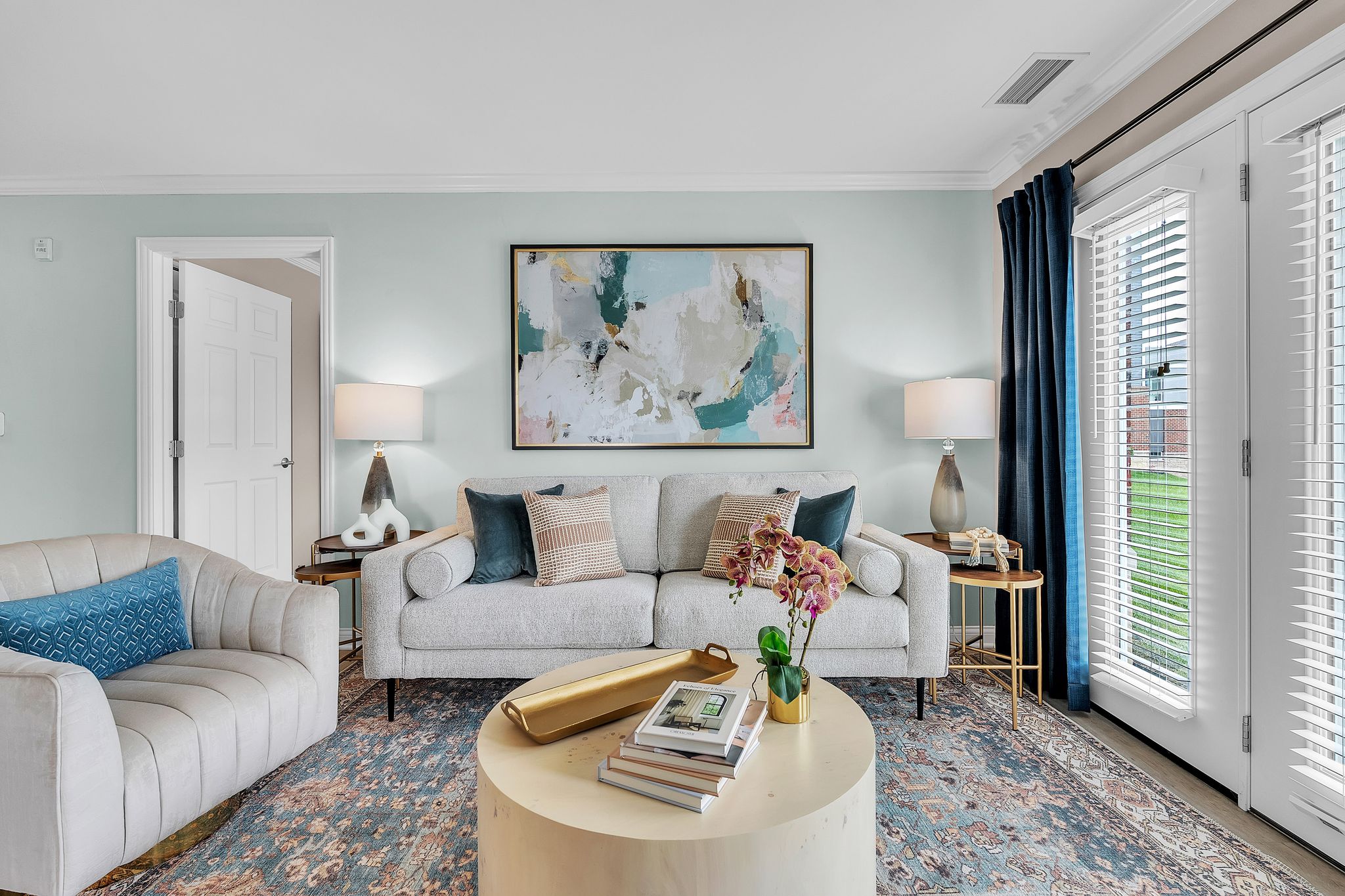 A modern, luxury living room in Hilliard Ohio features a light grey sofa, abstract art above it, two side tables with lamps, a round coffee table with books and flowers, an armchair, and large windows with dark curtains letting in natural light.