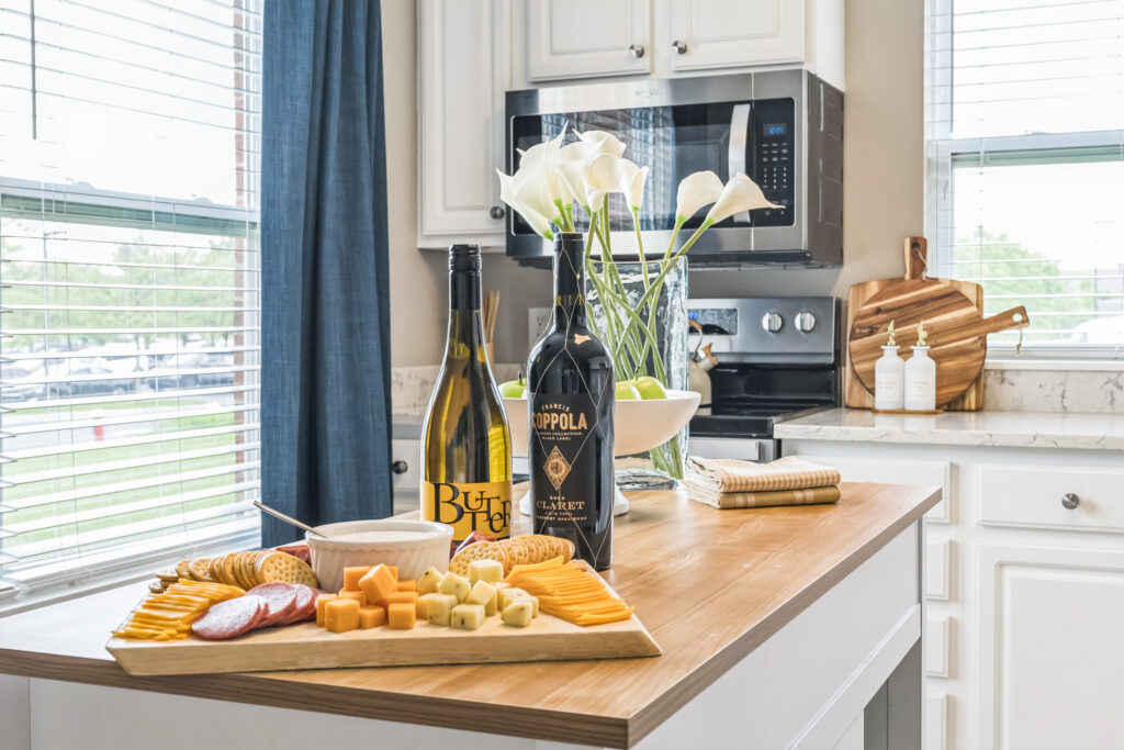 A luxury kitchen in Hilliard, Ohio features a wooden island with a cheese and cracker platter, two wine bottles, and a white bowl. White cabinets, a stove, and calla lilies in a vase complete this independent living space.
