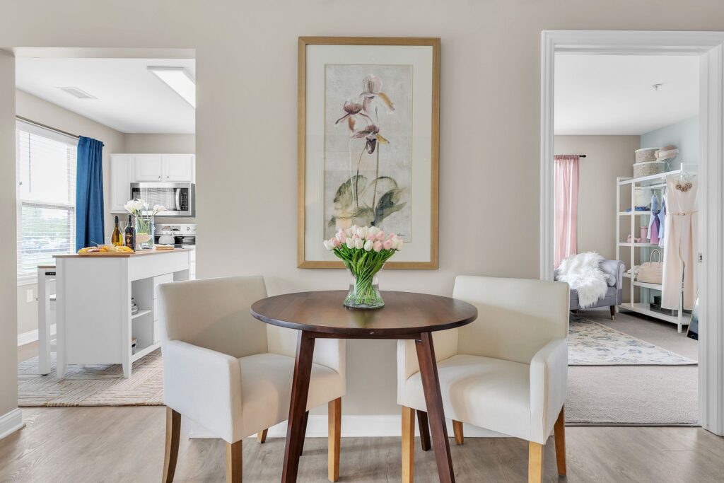 A round wooden table with two white upholstered chairs, a vase of pink tulips, and a framed flower painting on the wall. Enjoy luxury independent living in Hilliard Ohio, with doorways revealing a bright kitchen and cozy bedroom.