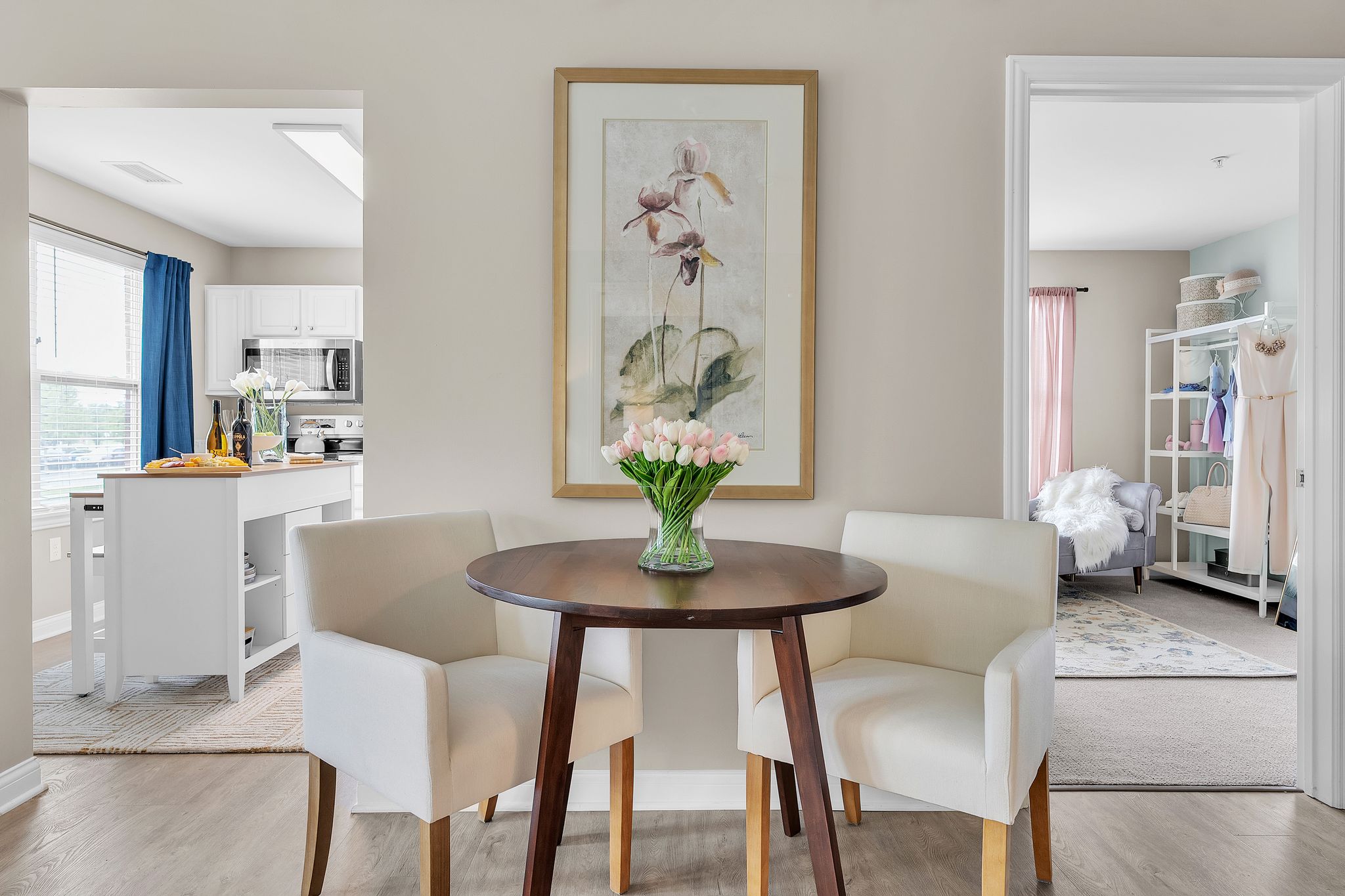 A round wooden table with two white upholstered chairs, a vase of pink tulips, and a framed flower painting on the wall. Enjoy luxury independent living in Hilliard Ohio, with doorways revealing a bright kitchen and cozy bedroom.