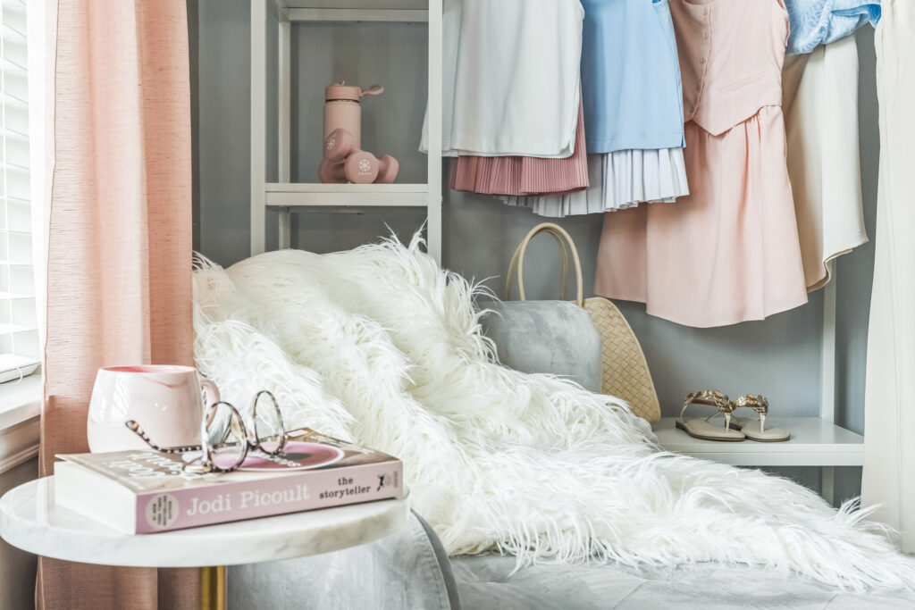 A cozy reading nook designed for independent living, featuring a fluffy white blanket on a chair, a round side table with a pink mug, eyeglasses, and a Jodi Picoult book. Pastel clothes and shoes are neatly arranged on nearby shelves.
