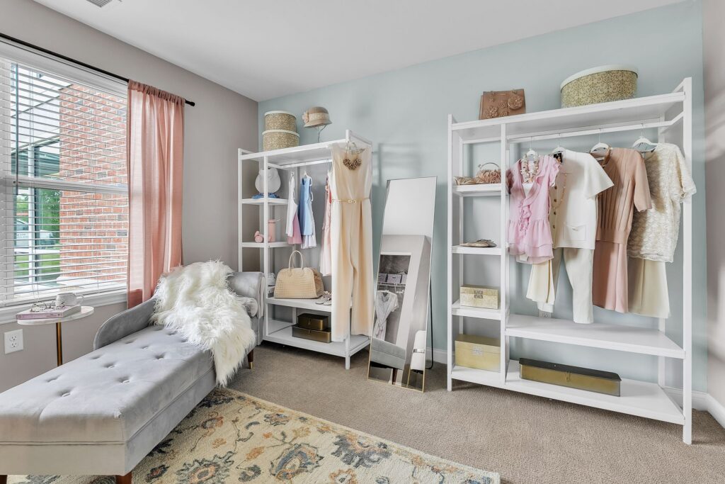 A stylish, luxury dressing room in Hilliard, Ohio features a gray chaise lounge, white open wardrobes with pastel dresses and handbags, a large mirror, and a window with pink curtains letting in natural light. Perfect for any senior’s elegant retreat.