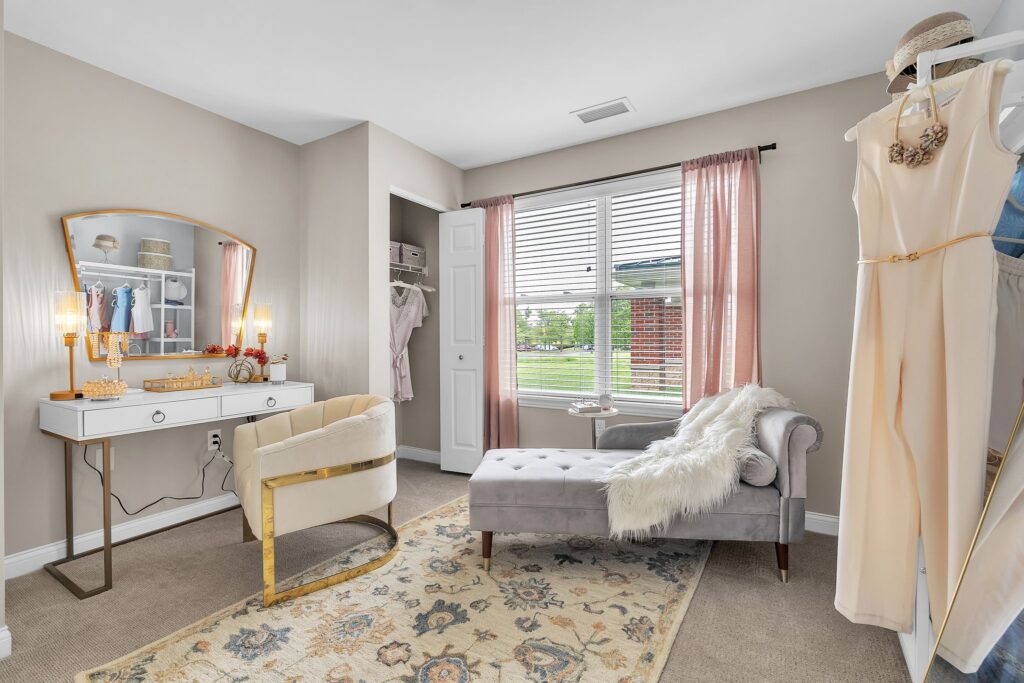 Bright, stylish dressing room with a vanity table, chair, and chaise lounge on a patterned rug. Pink curtains frame a large window, while neutral decor offers luxury and comfort—perfect for senior living or anyone seeking a cozy atmosphere.