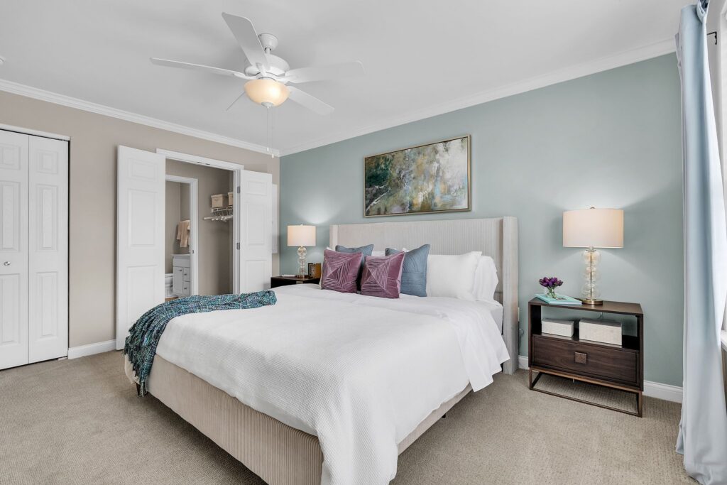 Experience luxury in this modern Hilliard, Ohio bedroom featuring a beige bed, plush bedding, colorful accent pillows, elegant nightstands with lamps, a ceiling fan, and walk-in closet—perfect for independent living.