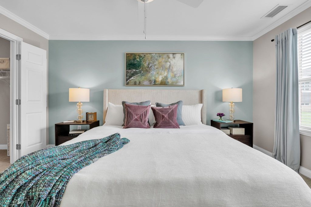 A modern bedroom designed for senior independent living in Hilliard, Ohio, featuring a neatly made bed with purple pillows, a blue-green throw, bedside tables with lamps, a landscape painting above the headboard, and sunlit curtains.