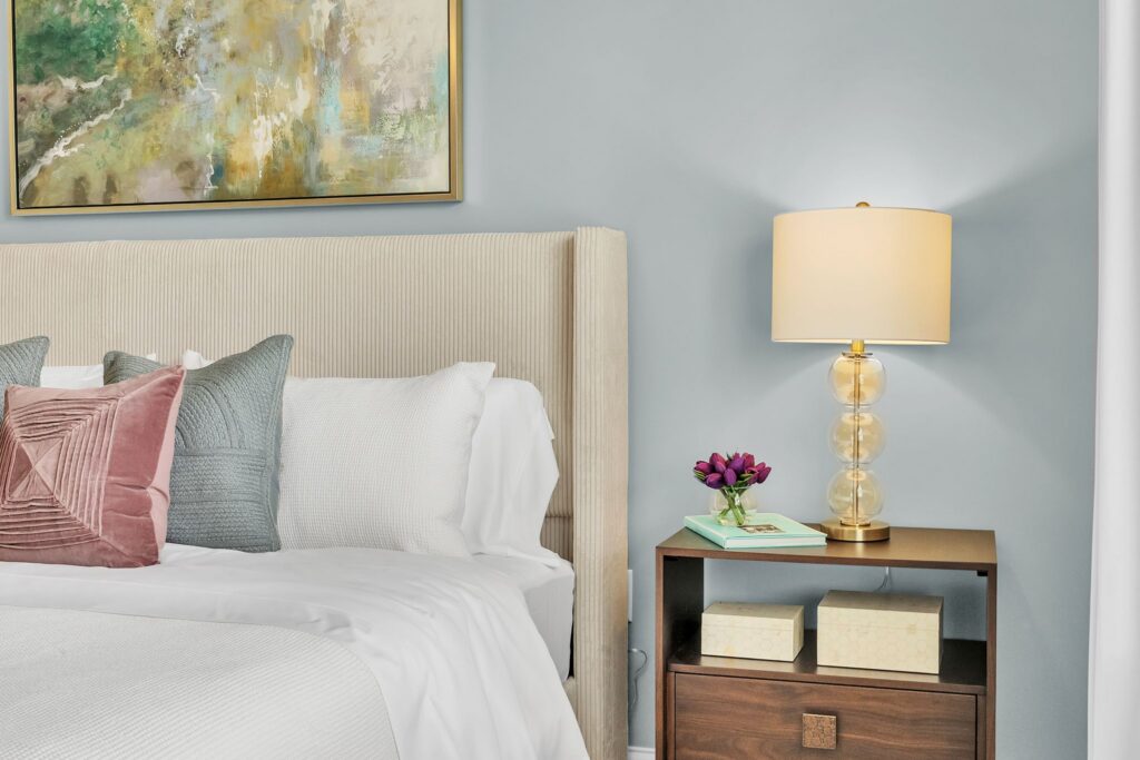 A modern bedroom designed for independent living in Hilliard, Ohio features a beige upholstered bed with white bedding and pastel pillows. A wood nightstand holds a glass lamp, purple flowers, books, and decorative boxes beneath an abstract painting.
