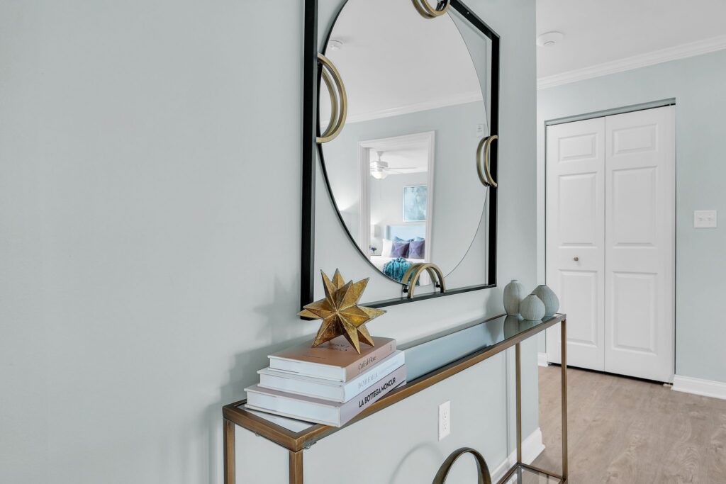 A modern hallway in this luxury residence features a glass console table with stacked books, a gold star sculpture, and two decorative vases. A large round mirror with a square frame hangs above, reflecting a bright living space.