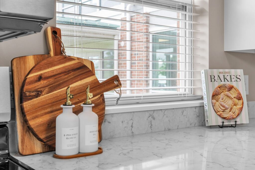 A kitchen countertop in an independent living space features two wooden cutting boards, oil dispensers, and a cookbook titled BAKES on a stand. A window with blinds offers an exterior view, perfect for senior residents who love to cook.