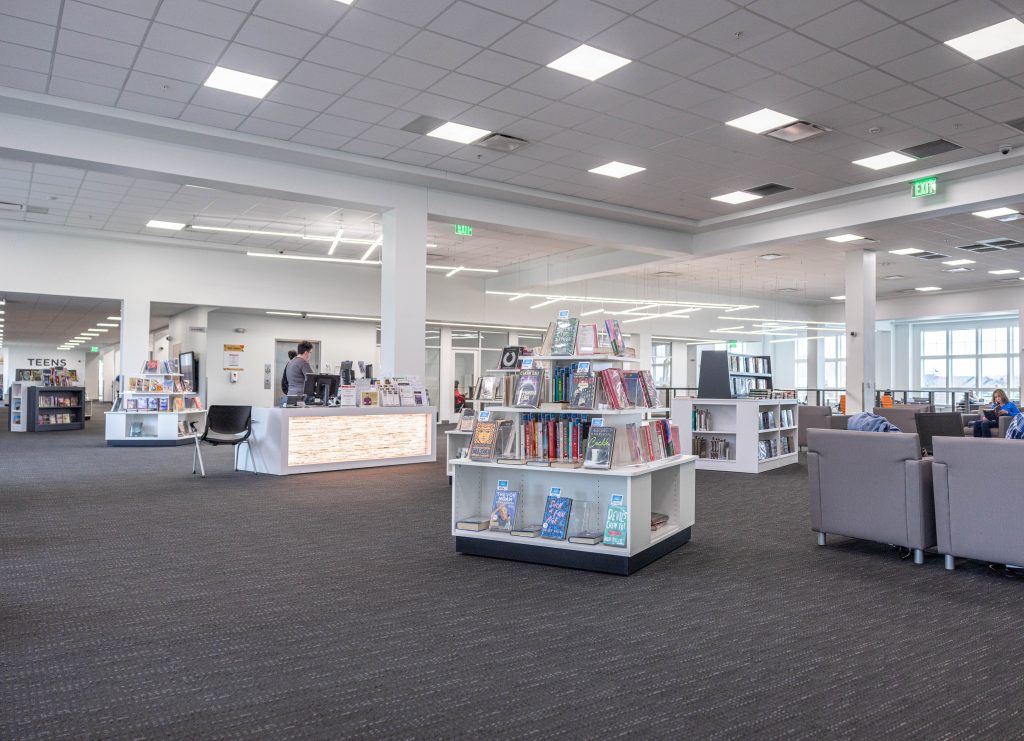 A modern library interior in Hilliard, Ohio, features bookshelves, a central display of books, a reception desk, a senior standing nearby, and a seating area with gray chairs. Bright lighting and large windows create an open, airy space for independent living.