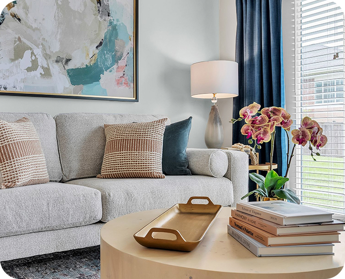 A modern living room designed for senior living, featuring a light gray sofa, patterned pillows, abstract wall art, a table lamp, and a round wooden coffee table with books, a gold tray, and a potted orchid near blue-curtained windows.