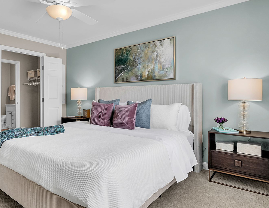 A modern bedroom with a large bed, white bedding, decorative pillows, and bedside tables exudes luxury. A painting above the headboard and an open door to the bathroom complete this stylish Hilliard Ohio retreat.