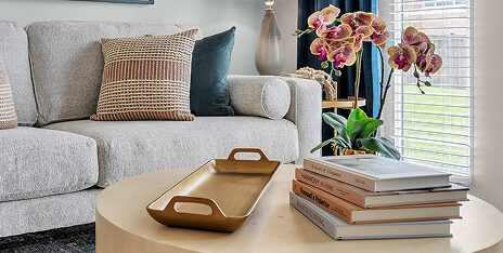 A beige couch with patterned pillows sits behind a round wooden coffee table holding a golden tray, a stack of books, and a potted orchid—creating a cozy space ideal for independent living in Hilliard Ohio, near a window with natural light.