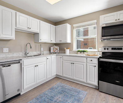 A modern kitchen with white cabinets, stainless steel appliances, a dishwasher, an electric stove, a large window, and a light blue rug on a wood floor. Countertops are light-colored stone.