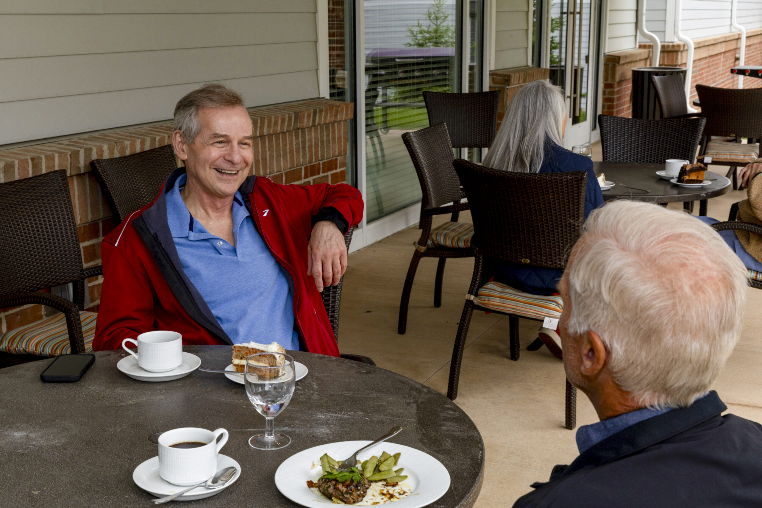 Two senior men sit at an outdoor café table in Hilliard Ohio, smiling and chatting over coffee and dessert. Another couple sits in the background, enhancing the relaxed, friendly atmosphere perfect for senior living.