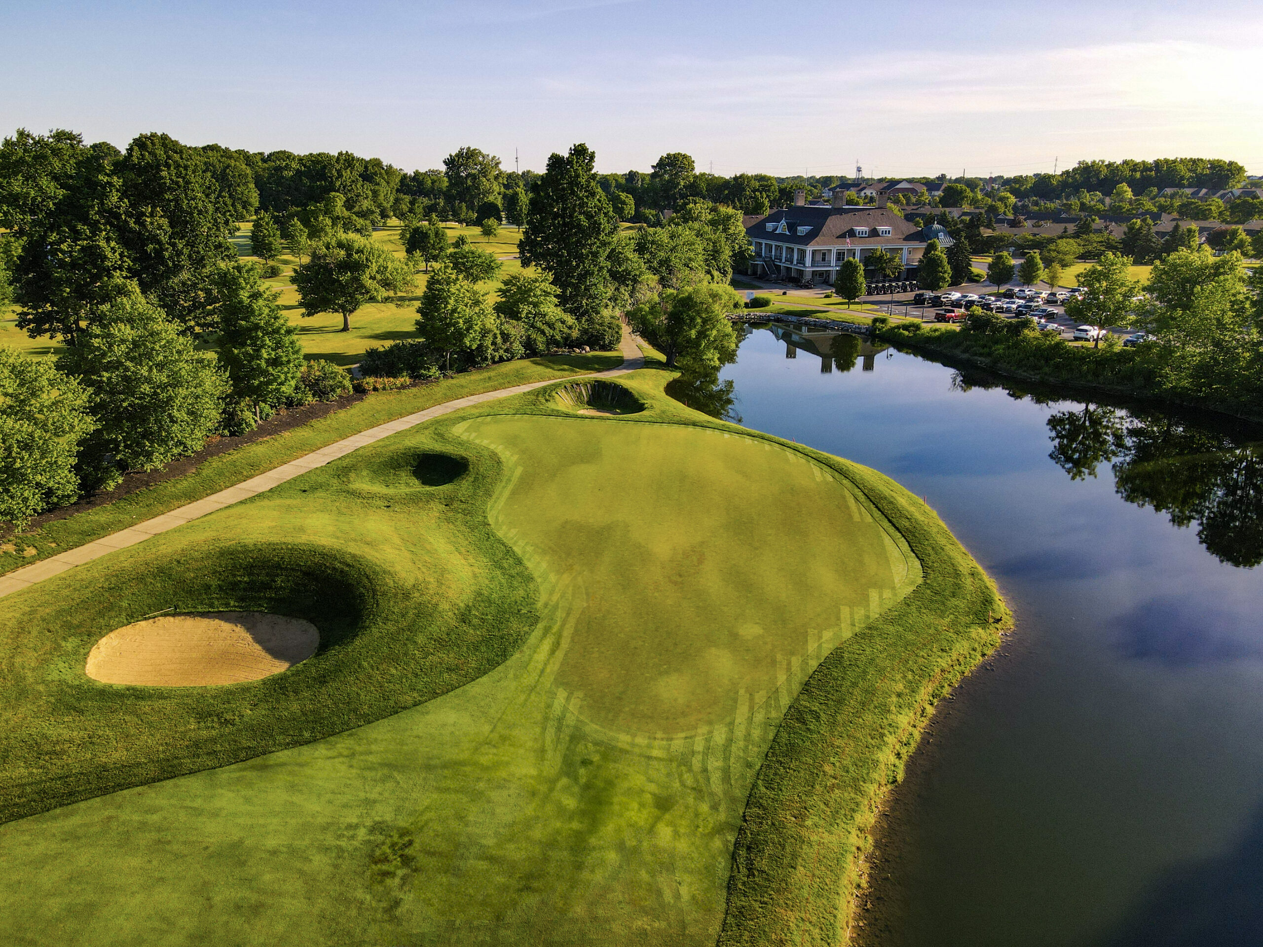 Aerial view of a golf course green in Hilliard, Ohio bordered by sand bunkers and a pond, with a clubhouse, parked cars, and trees—an ideal spot for independent living under a clear blue sky.