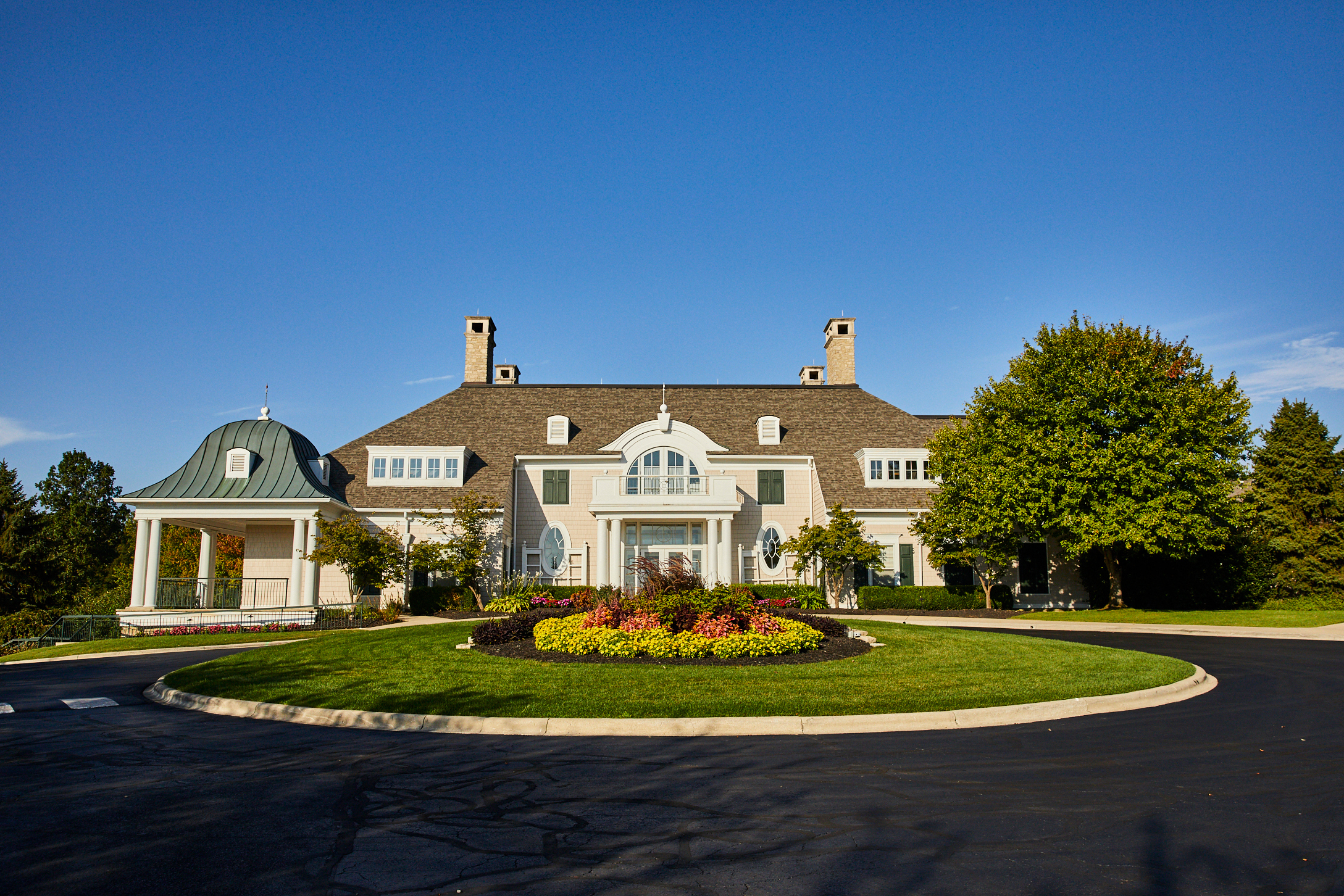 A large, elegant mansion in Hilliard with white walls, tall chimneys, and a manicured circular driveway bordered by colorful flowers and lush green trees offers luxury senior living under a clear blue sky.