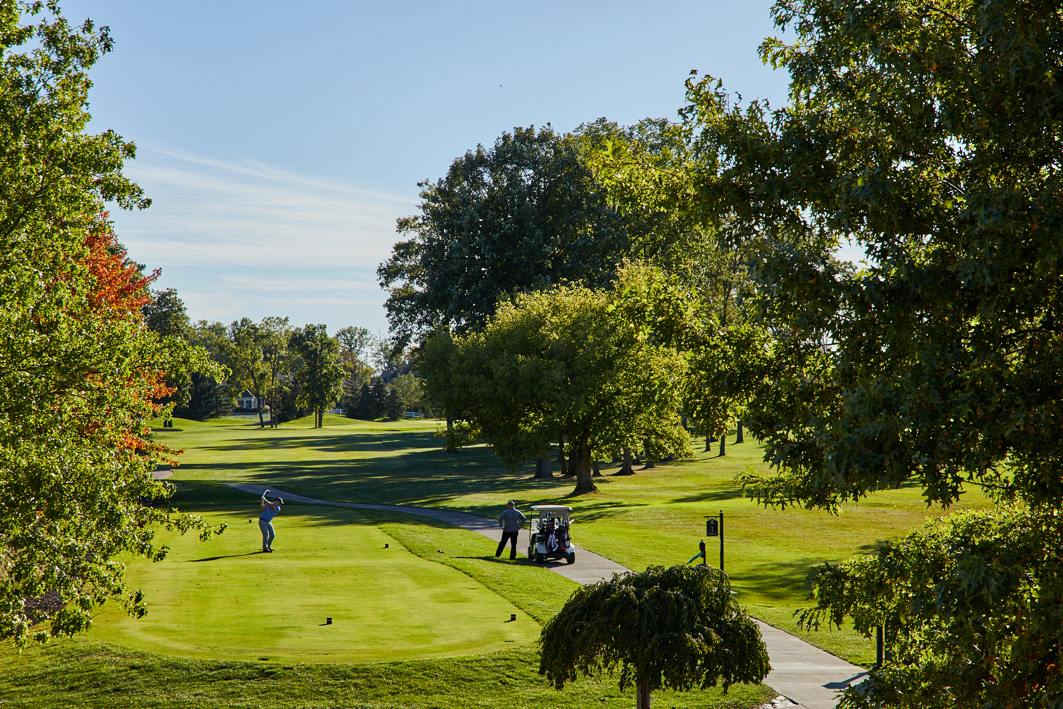 Two people enjoy a round of golf on a green Ohio course surrounded by large trees under a clear blue sky; one person swings a club while the other stands by a golf cart, showcasing the active lifestyle in senior living.