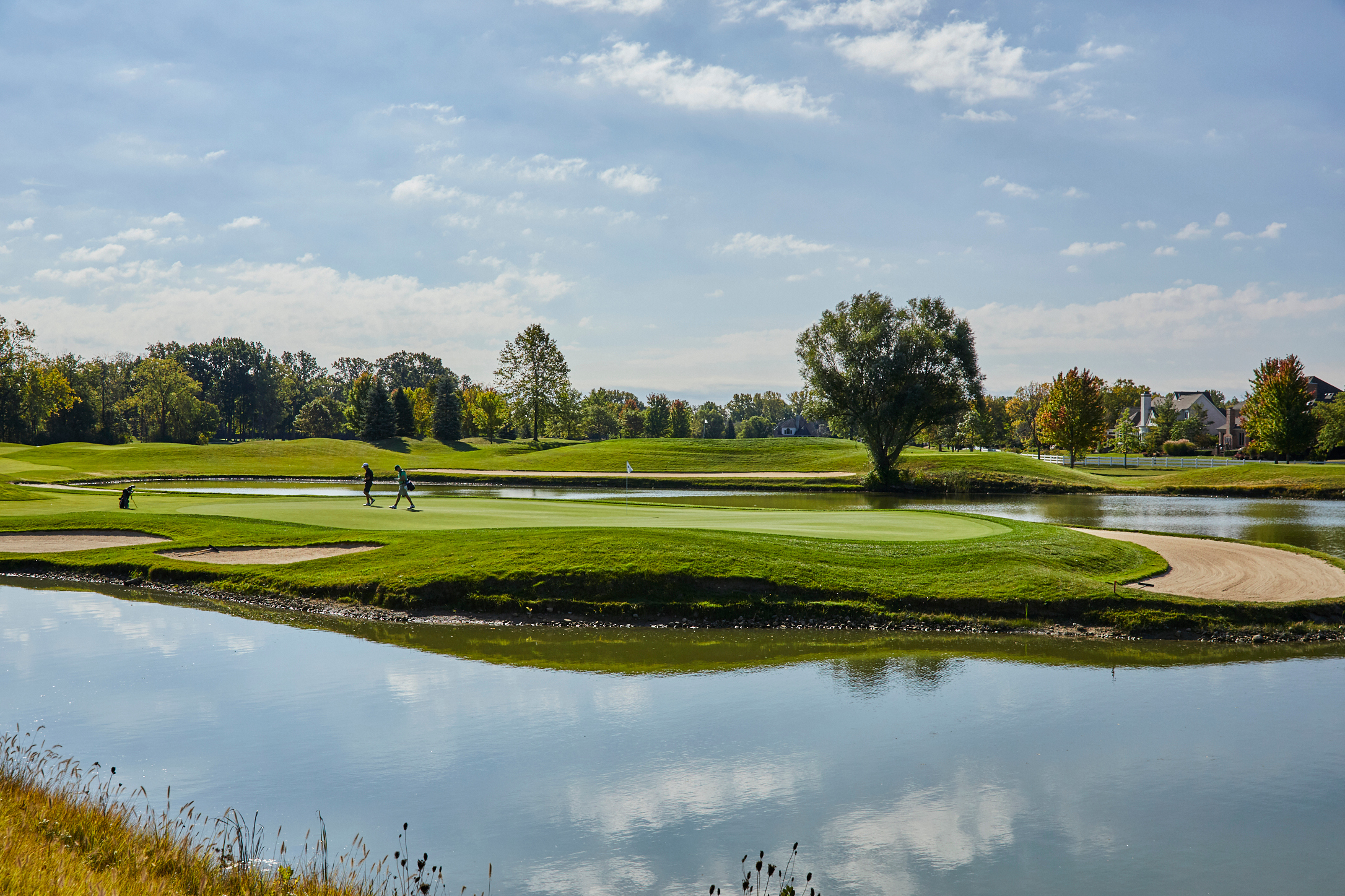 Two people walk across a green golf course in Hilliard, surrounded by a calm pond, sand bunkers, and trees under a partly cloudy sky—perfect for seniors enjoying independent living. The landscape is reflected in the water.