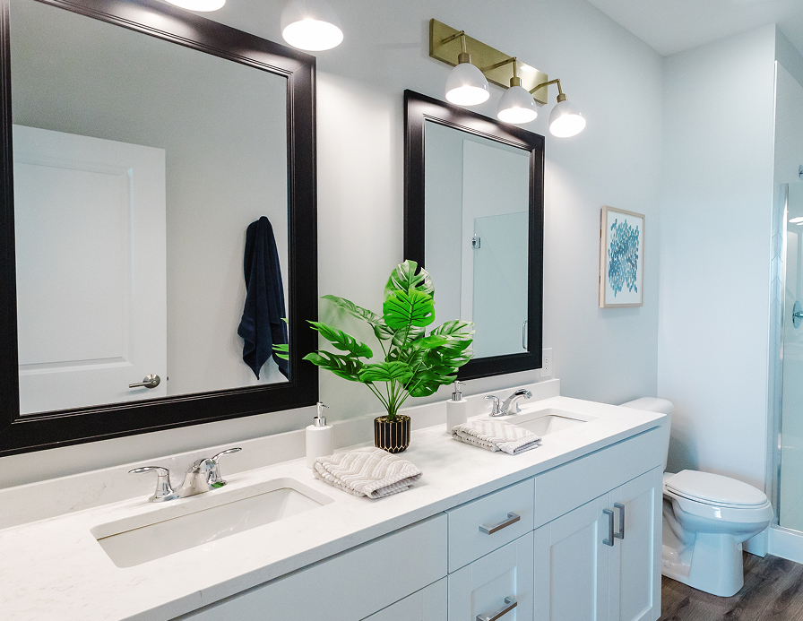 Modern bathroom with double sinks, large black-framed mirrors, a potted green plant between the sinks, a set of towels, wall-mounted lights, and luxury touches for comfortable independent living; toilet and shower visible in the background.