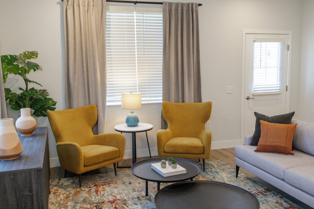 A modern living room in a Hilliard Ohio senior living community features two yellow armchairs, a small round table with a lamp, a gray sofa with orange pillows, a patterned rug, houseplants, and light gray curtains over the window.