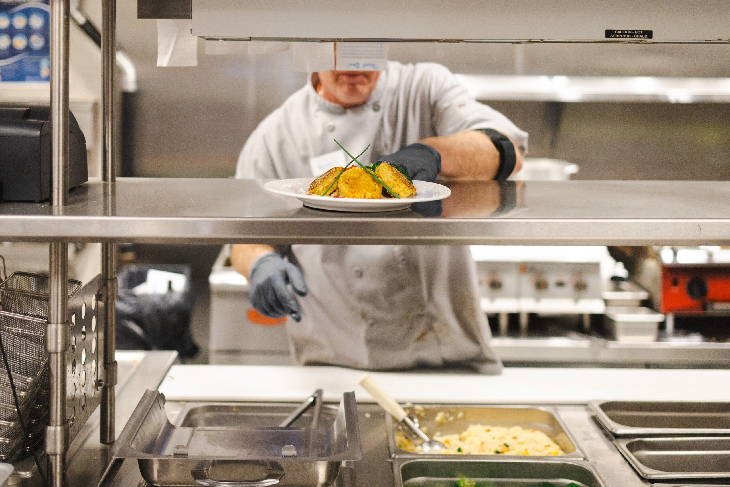 A chef in a white uniform and gloves places a luxury plated dish garnished with herbs on a metal counter in a commercial kitchen, showcasing the elevated dining experience offered in senior independent living communities.