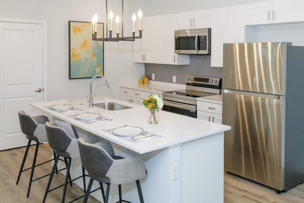 Experience luxury in this modern Hilliard, Ohio kitchen, featuring white cabinets, stainless steel appliances, a spacious island with seating—perfect for senior living and entertaining beneath a stunning chandelier and colorful abstract painting.