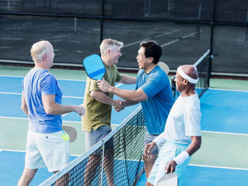Four senior men stand on a pickleball court in Hilliard, Ohio, smiling and shaking hands across the net after a game. Dressed in athletic wear and holding paddles, they enjoy a friendly match in a luxury setting.