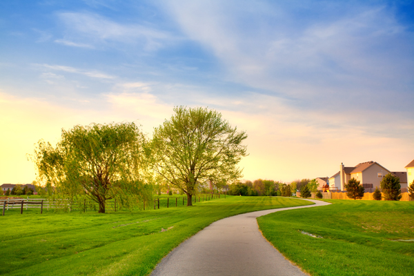 A paved sidewalk curves through a green, grassy park in Hilliard, Ohio, with trees on the left and luxury houses on the right, under a blue sky with soft clouds at sunset.