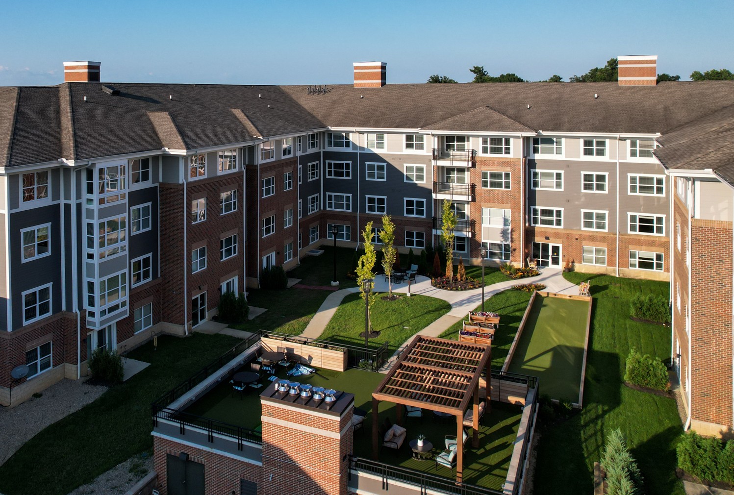 Aerial view of a modern, three-story apartment building with a central courtyard featuring green lawns, walking paths, seating areas, a pergola, and a bocce court under clear blue skies.
