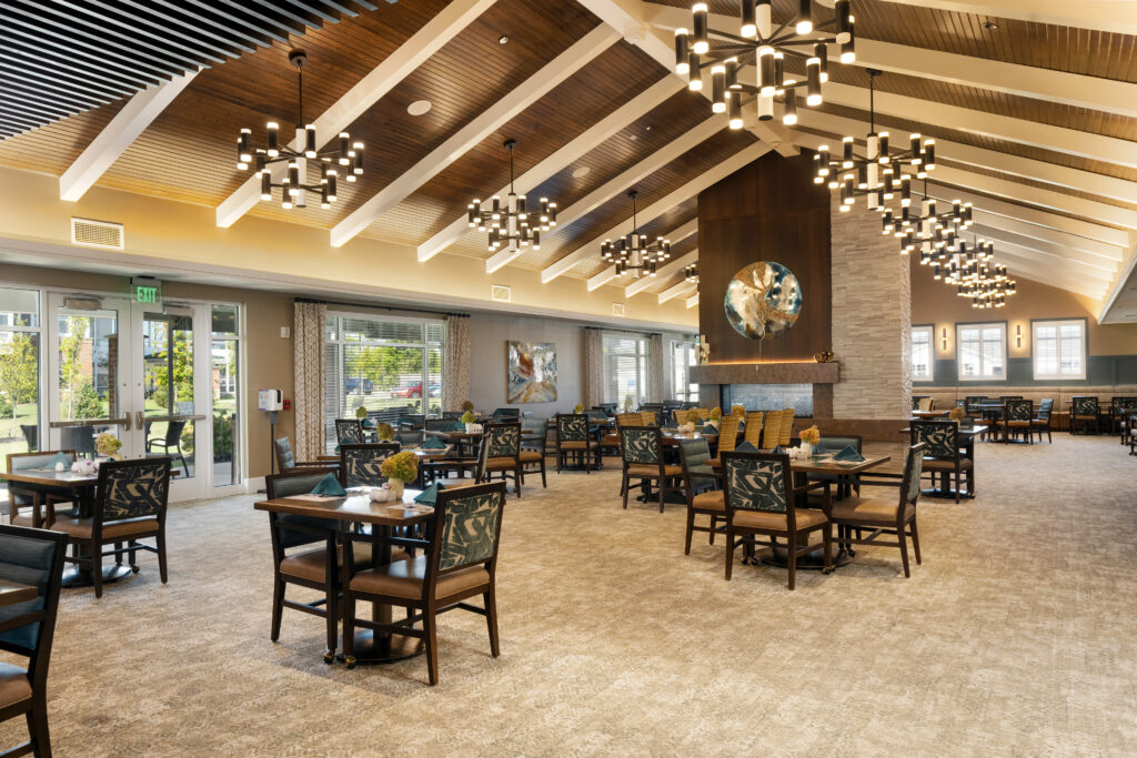 A spacious, elegant dining area designed for luxury independent living, featuring multiple tables and chairs, warm chandelier lighting, high vaulted ceilings with wooden beams, large windows, and a central fireplace adorned with a circular mirror.