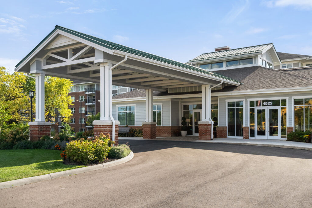 A modern luxury building with a covered driveway entrance, large windows, and landscaped gardens. The exterior features white pillars, brick accents, and a green roof—ideal for independent living at address 4522 near the entrance.