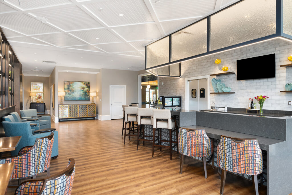 Modern lounge area designed for luxury independent living, featuring a bar, high chairs, and patterned armchairs. Wood flooring and contemporary decor in neutral tones with pops of color create a welcoming space for seniors. A large painting and shelves adorn the background.