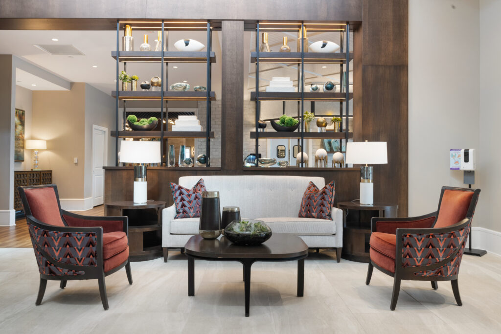 Modern lobby in Hilliard, Ohio, featuring a beige sofa, two patterned armchairs, a round black coffee table with a glass bowl, and open shelving for books and decor—perfect for independent living or senior living communities.