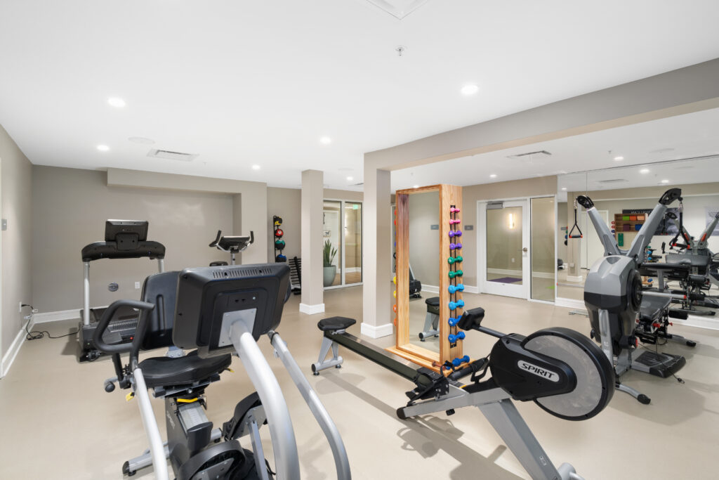 A modern home gym designed for senior living, featuring exercise equipment like bikes, treadmills, a rowing machine, and free weights on a stand—set in a bright, well-lit room with light-colored flooring and wall mirrors.