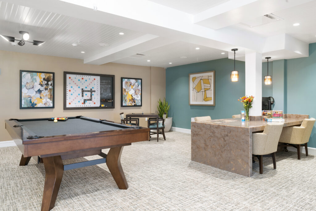 A modern recreation room in this luxury senior living community features a pool table, bar area, stylish seating, wall art, and pendant lights. Neutral walls and carpet create a warm ambiance, enhanced by vibrant plants and flowers.