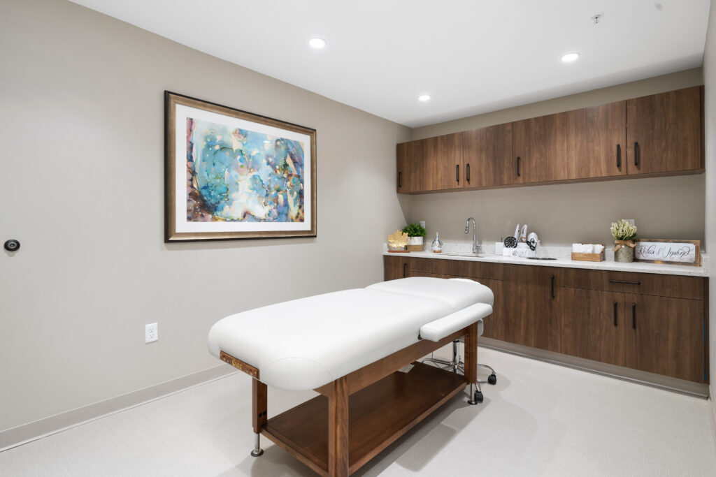 A modern luxury spa treatment room with a white massage table, wooden cabinets, a countertop with a sink, and abstract artwork on the wall under bright lighting. The space is clean, organized, and perfect for senior living wellness.