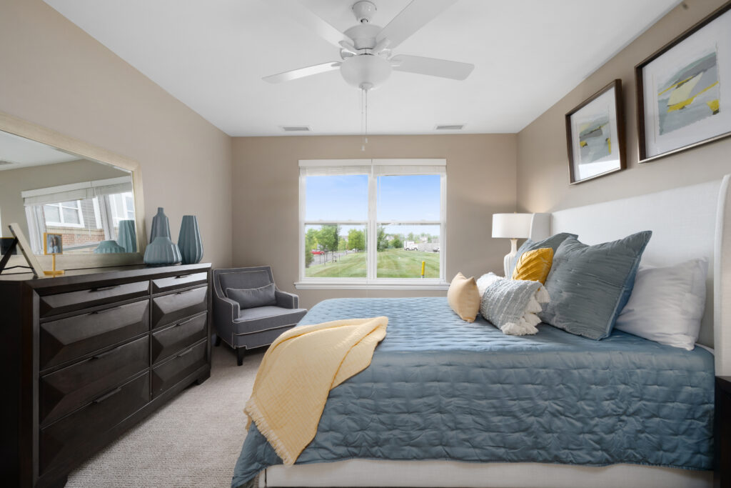 A bright luxury bedroom with a bed covered in blue and yellow bedding, a dark wooden dresser with decorative vases, an armchair—ideal for senior living—and artwork on the wall beside a large window flooding the space with natural light.