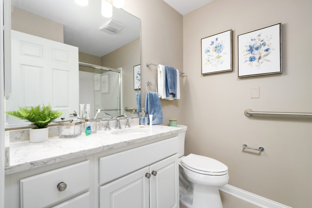 Modern bathroom with white cabinets, a marble countertop, sink, toilet, folded blue and white towels, two floral wall art pieces, a potted plant, and safety grab bars on beige walls—perfect for luxury senior living.