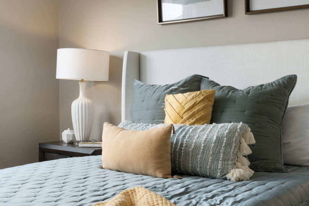A neatly made bed with blue and beige pillows sits next to a dark nightstand with a white table lamp and small decorative items, creating a cozy, luxury feel ideal for senior independent living. Light walls and framed artwork complete the inviting space.