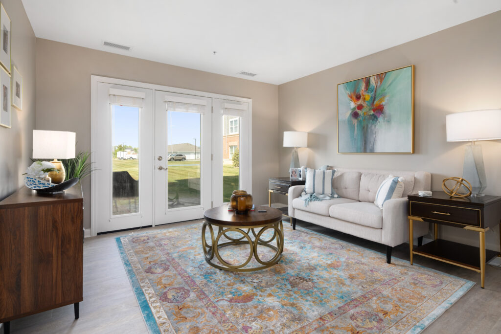Bright living room with a beige sofa, colorful rug, round wooden coffee table, large artwork above the couch, two side tables with lamps, and double glass doors letting in natural light.
