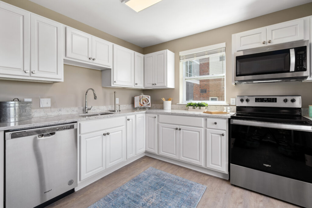 A bright, modern kitchen designed for independent living features white cabinets, stainless steel appliances, marble countertops, and a window filling the space with natural light. A blue rug accents the light wood floor.