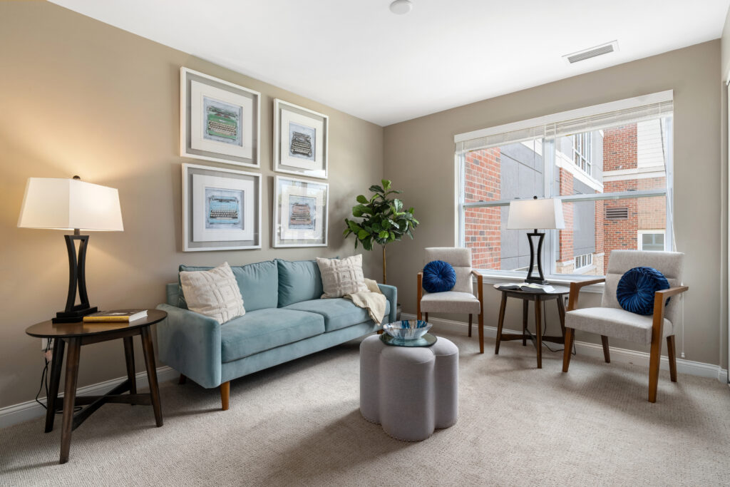 A modern living room in Hilliard, Ohio with a light blue sofa, two white armchairs with blue pillows, a round side table, floor lamp, wall art, large window with blinds, and a potted plant—perfect for comfortable senior living.