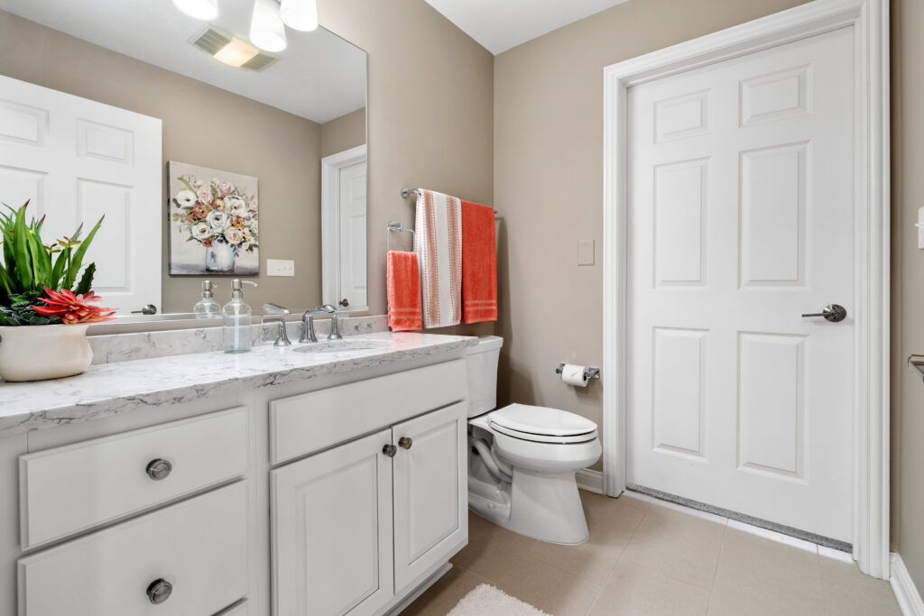 A clean, modern bathroom in Hilliard, Ohio features a white vanity, marble countertop, sink, toilet, large mirror, beige walls, floral wall art, potted plant, and orange towels—perfect for comfortable independent living.