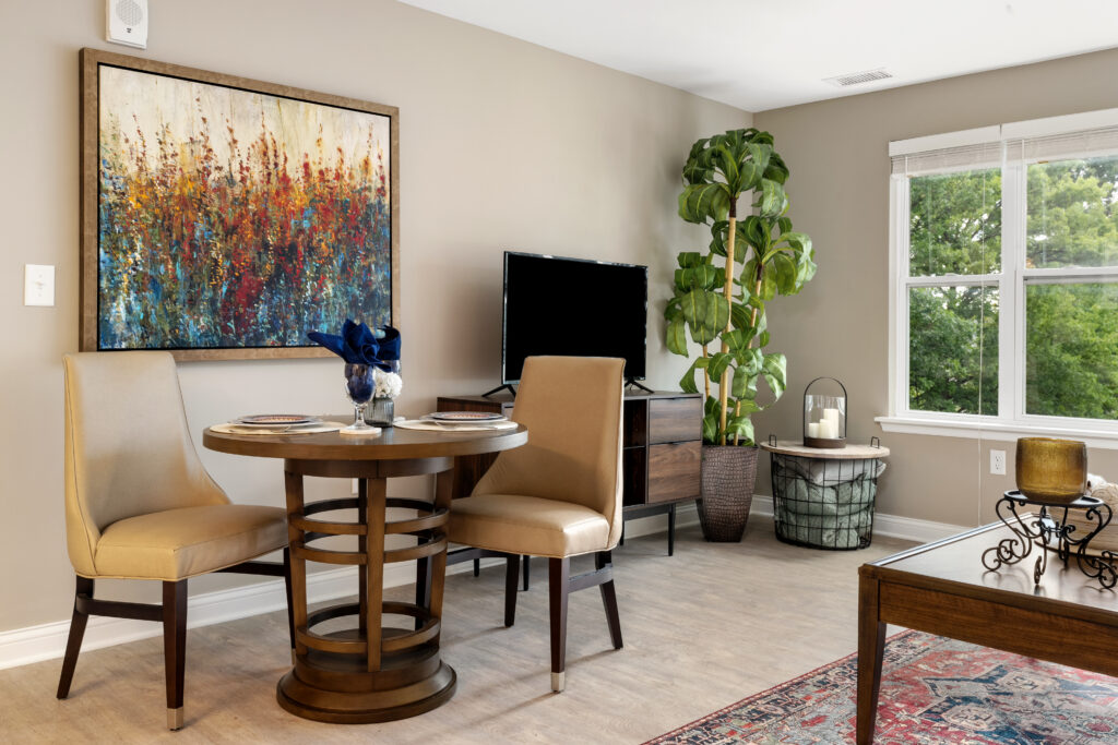 A modern living space in Hilliard, Ohio, featuring a round wooden table with two beige chairs, a TV on a stand, luxury décor touches, a large potted plant, and a window overlooking green trees—perfect for independent living.