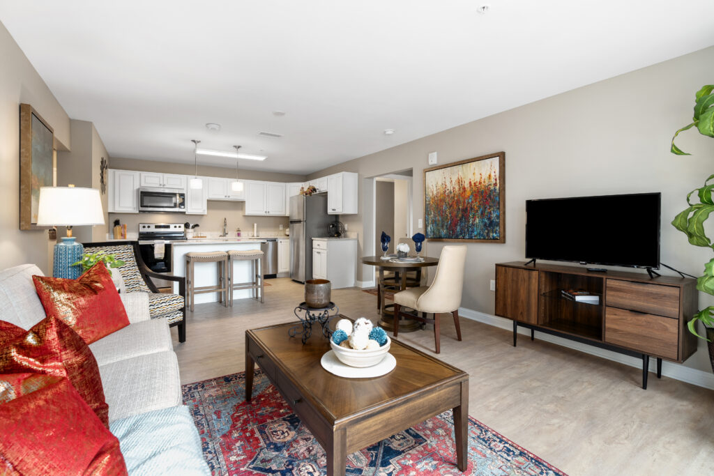 A modern, open-concept living room and kitchen in Hilliard Ohio, featuring neutral walls, a gray sofa with red pillows, a wooden coffee table, TV console, dining area, and white luxury kitchen cabinets with stainless steel appliances.