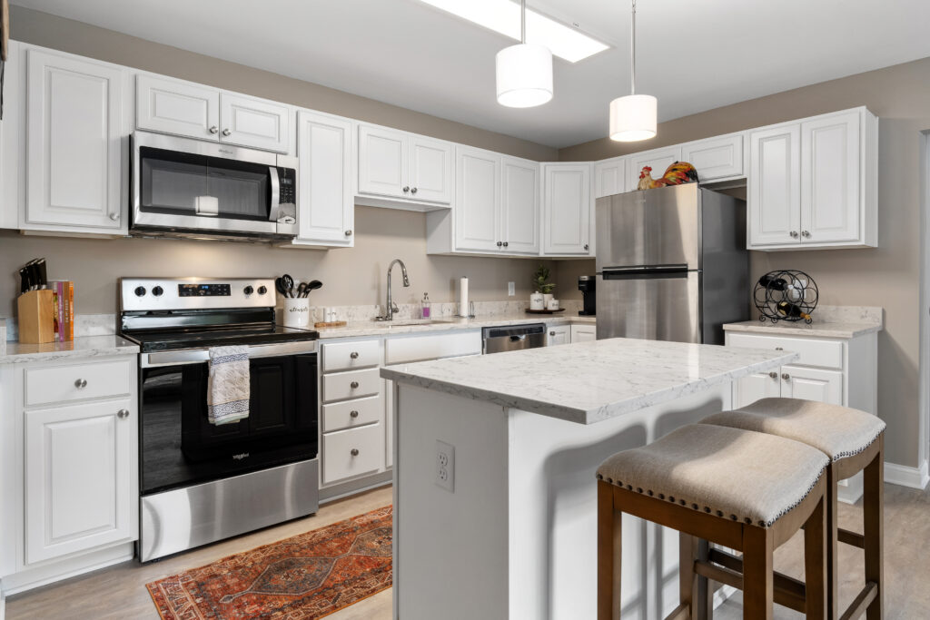 Modern kitchen with white cabinets, stainless steel appliances, marble countertops, and a central island with two cushioned stools—perfect for luxury independent living in Hilliard Ohio. Pendant lighting and a decorative rug add warmth to the space.