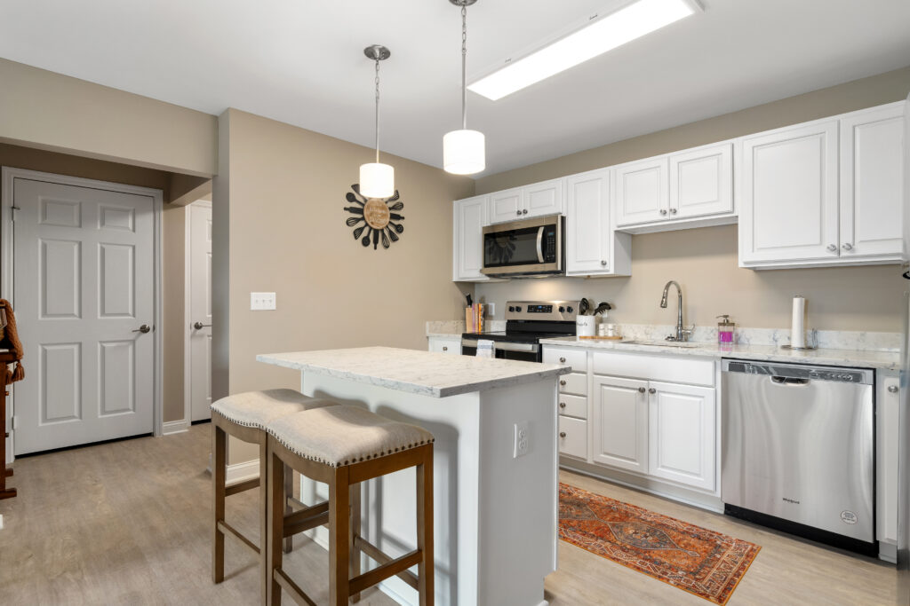Modern kitchen with white cabinets, stainless steel appliances, a small island with two upholstered stools—perfect for luxury senior living. Light wood flooring, beige walls, and stylish decor create an inviting, independent living space.