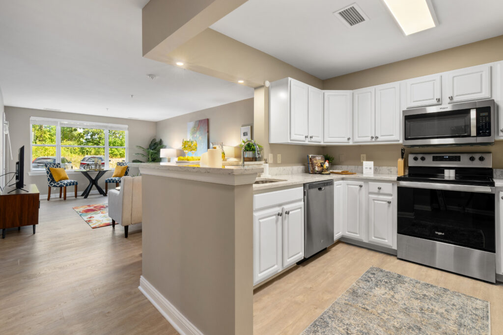 Modern open-concept kitchen and living area with white cabinets, stainless steel appliances, light wood flooring, and large windows for natural light—perfect for luxury senior living. Dining and sitting areas are visible in the background.